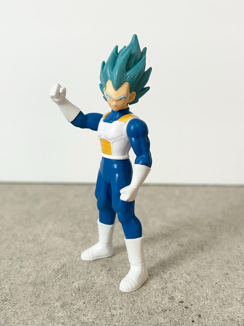 Pode incluir: Uma figura de a&ccedil;&atilde;o de pl&aacute;stico de um personagem com cabelo espetado azul-petr&oacute;leo, vestindo um traje azul com armadura de peito branca e amarela, luvas brancas e botas brancas, em uma pose com um bra&ccedil;o levantado e o punho fechado.