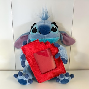 May include: A plush toy of the character Stitch from Lilo & Stitch, in shades of blue, with large ears and holding a red photo frame. The toy has a tuft of blue hair and is sitting on a white surface.