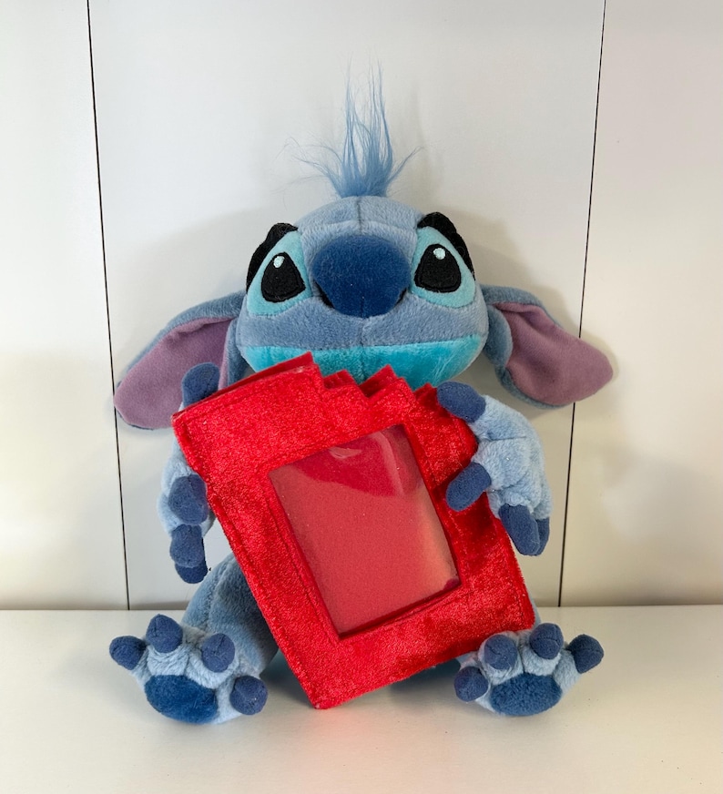 May include: A plush toy of the character Stitch from Lilo & Stitch, in shades of blue, with large ears and holding a red photo frame. The toy has a tuft of blue hair and is sitting on a white surface.
