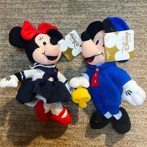 May include: Two Disney Store plush toys: Minnie Mouse in a sailor outfit with a red bow and shoes, and Mickey Mouse in a blue jacket and hat. Both have tags that say "The Disney Store".