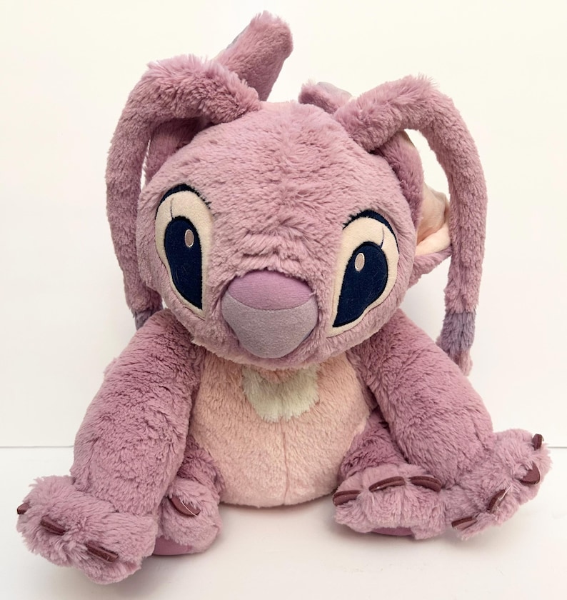 May include: A plush toy of the character Angel from the Lilo & Stitch franchise. The toy is primarily lavender with a light pink chest and inner ears. It has large, dark blue eyes and long, floppy ears.