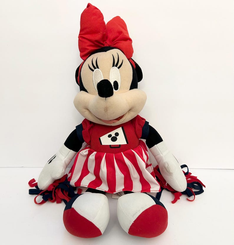 May include: A plush Minnie Mouse doll wearing a red and white cheerleader outfit with a matching bow. The doll has black ears, a black nose, and white gloves. The outfit includes a red top with a white and black graphic and a red and white striped skirt.