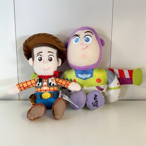 May include: Two plush toys of the characters Woody and Buzz Lightyear from the movie Toy Story. Woody is wearing a cowboy hat, plaid shirt, and blue jeans. Buzz Lightyear has a purple head, green and white spacesuit, and the word "Andy" on his foot.
