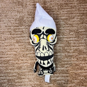 May include: A plush toy in the shape of a skeleton with a white nightcap. The toy has a cream-colored face with black markings, large yellow eyes, and a wide grin. The toy is lying on a brown carpet.