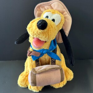 May include: A plush yellow dog toy with long black ears, a pink tongue, and white eyes. The dog wears a tan explorer hat, a bright blue neckerchief, and a brown satchel across its chest. The toy has a soft, fuzzy texture throughout its body.