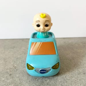 May include: A light blue toy car featuring a cartoon character with a small yellow curl on its head emerging from the roof. The vehicle has an orange windshield, yellow headlights, and a silver front grille with the VTech logo printed in blue.