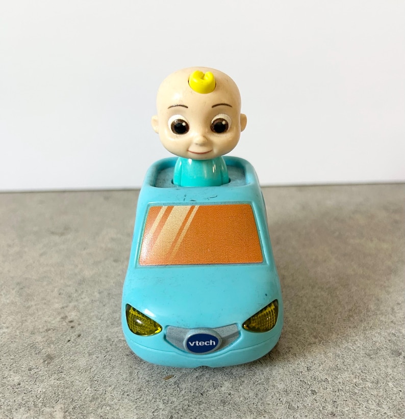 May include: A light blue toy car featuring a cartoon character with a small yellow curl on its head emerging from the roof. The vehicle has an orange windshield, yellow headlights, and a silver front grille with the VTech logo printed in blue.