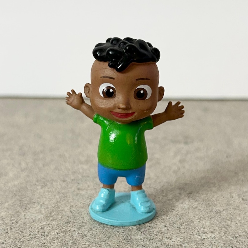 May include: A small plastic figurine of a child with dark skin and black hair, wearing a bright green t-shirt and blue shorts. The figure has large brown eyes, a smiling mouth, and is standing on a round blue base with arms outstretched to the sides.