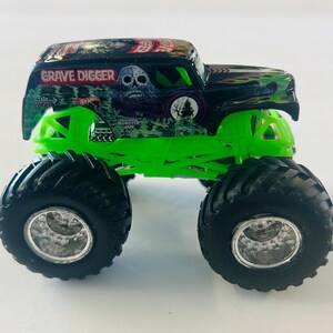 May include: A Hot Wheels Grave Digger monster truck toy. The truck has a black and green body with the words "Grave Digger" and a skull graphic. It features large black tires and bright green suspension.
