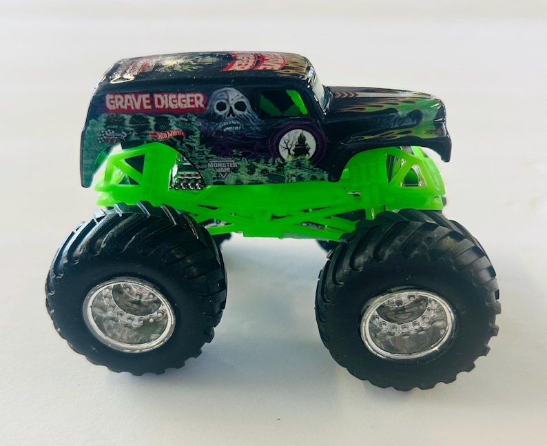 May include: A Hot Wheels Grave Digger monster truck toy. The truck has a black and green body with the words "Grave Digger" and a skull graphic. It features large black tires and bright green suspension.