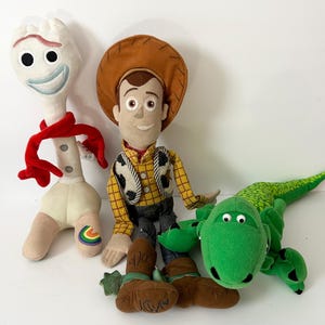 May include: Three plush toys including a white spork character with a red scarf, a cowboy doll wearing a brown hat and yellow plaid shirt, and a green dinosaur with a textured back. The cowboy doll has the name Andy written on its brown boots.