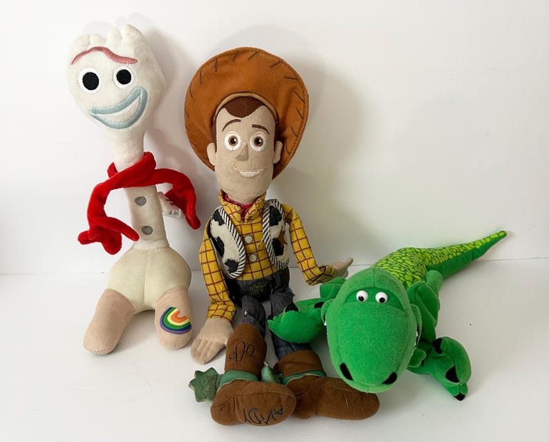 May include: Three plush toys including a white spork character with a red scarf, a cowboy doll wearing a brown hat and yellow plaid shirt, and a green dinosaur with a textured back. The cowboy doll has the name Andy written on its brown boots.