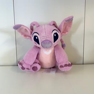 May include: A pink plush toy of the character Angel from Lilo & Stitch. The toy has large ears, big black eyes, and a white heart-shaped design on its chest. The toy is sitting on a white surface.