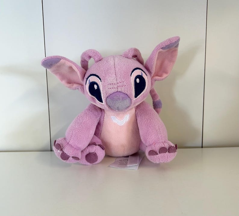 May include: A pink plush toy of the character Angel from Lilo & Stitch. The toy has large ears, big black eyes, and a white heart-shaped design on its chest. The toy is sitting on a white surface.