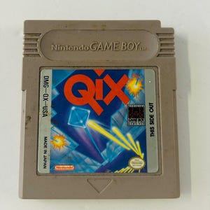 May include: A Nintendo Game Boy cartridge for the game Qix. The cartridge is a light gray color. The label features the word "QIX" in large red letters, with a blue and purple background. The cartridge also has the text "MADE IN JAPAN".