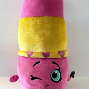 May include: A plush toy shaped like a lipstick tube, primarily pink with a yellow band and crown. The toy has a winking face with a large blue eye and small arms and feet. It is a whimsical, colorful design.