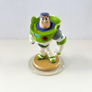 Puede incluir: Una figura de Buzz Lightyear de la franquicia Toy Story. La figura es predominantemente blanca, verde y morada, con una cara sonriente y una pose decidida. Se encuentra sobre una base de color madera con un anillo de pl&aacute;stico transparente.