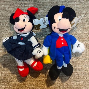 May include: Two plush Disney character dolls, Mickey Mouse and Minnie Mouse. Mickey wears a blue suit and red shirt. Minnie wears a navy dress and red shoes. Both dolls have tags attached.