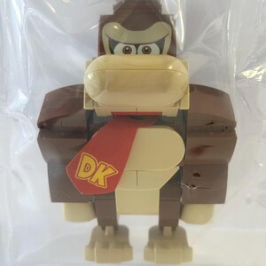 May include: A Donkey Kong figure constructed from interlocking plastic bricks. The figure features a brown head and body, a tan face, and a red tie with "DK" printed on it. The figure is packaged in a clear plastic bag.