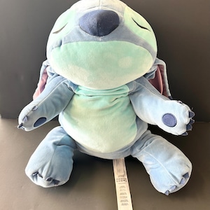 May include: A plush Stitch doll in shades of blue, teal, and purple, with closed eyes. The soft toy features large ears, a round body, and a tag. The doll is sitting on a gray surface.