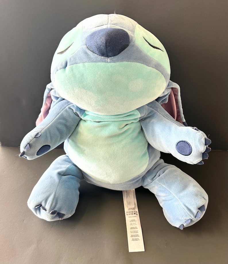 May include: A plush Stitch doll in shades of blue, teal, and purple, with closed eyes. The soft toy features large ears, a round body, and a tag. The doll is sitting on a gray surface.