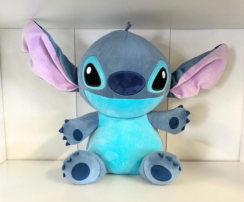 Może przedstawiać: Pluszowa zabawka postaci Stitch z filmu animowanego Lilo & Stitch. Zabawka jest gł&oacute;wnie niebieska z dużymi uszami, kt&oacute;re są r&oacute;żowe w środku. Ma duże czarne oczy i jasnoniebieski brzuch.