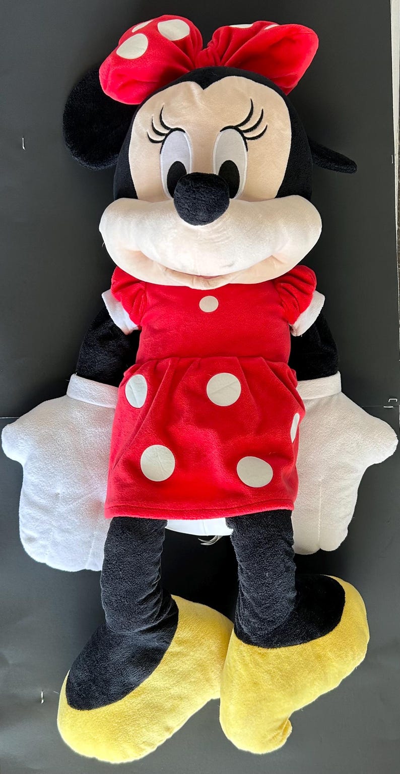 May include: A plush Minnie Mouse doll, featuring a red dress with white polka dots, a matching bow, and white gloves. The doll has black ears, a black nose, and yellow shoes. The toy is designed for children.