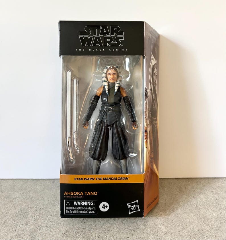 May include: A Star Wars The Black Series action figure of Ahsoka Tano in its original packaging. The figure wears a dark sleeveless tunic, black arm guards, and wide-legged pants. Two white lightsabers are included inside the clear plastic window.