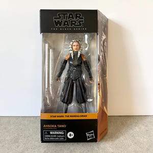 May include: A Star Wars The Black Series action figure of Ahsoka Tano in its original packaging. The figure wears a dark sleeveless tunic, black arm guards, and wide-legged pants. Two white lightsabers are included inside the clear plastic window.