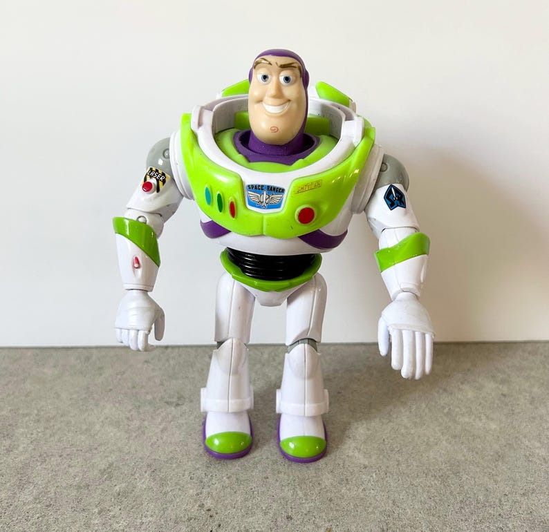 May include: A plastic action figure of a space ranger character with a purple hood, white and lime green armor, and a chest plate featuring the text SPACE RANGER. The figure has articulated limbs, a smiling face, and various colorful decals.