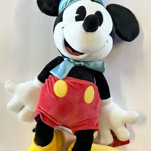 Pu&ograve; includere: Un peluche di Topolino con un cappello da festa blu e un papillon. Topolino ha orecchie nere, viso bianco e pantaloncini rossi con bottoni gialli. Le sue scarpe gialle e i guanti bianchi completano il look.