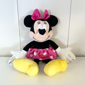 May include: A plush Minnie Mouse doll with a black body, pink skirt with white polka dots, and a pink bow. The doll has white gloves and yellow shoes. The doll is sitting upright.