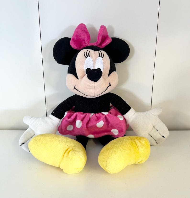 May include: A plush Minnie Mouse doll with a black body, pink skirt with white polka dots, and a pink bow. The doll has white gloves and yellow shoes. The doll is sitting upright.