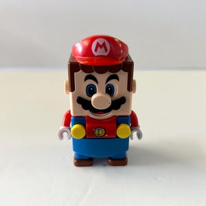 Puede incluir: Una figura de Lego de Mario, el ic&oacute;nico personaje de videojuegos. La figura tiene una gorra roja con una "M" blanca, una cabeza marr&oacute;n en forma de bloque y una camisa roja con un peto azul. La figura tiene un bigote negro y ojos azules.