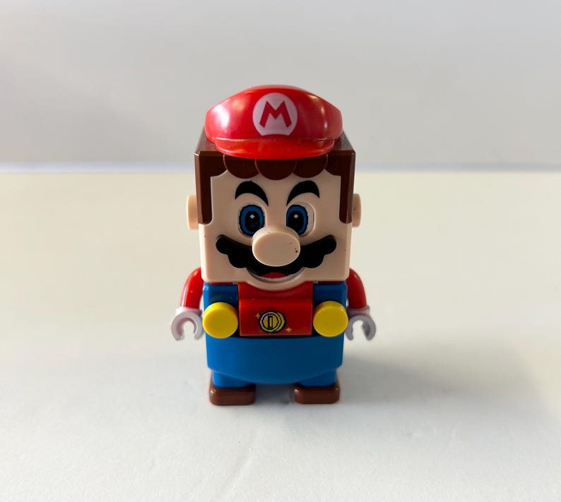Puede incluir: Una figura de Lego de Mario, el ic&oacute;nico personaje de videojuegos. La figura tiene una gorra roja con una "M" blanca, una cabeza marr&oacute;n en forma de bloque y una camisa roja con un peto azul. La figura tiene un bigote negro y ojos azules.