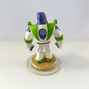 Puede incluir: Una figura de Buzz Lightyear, vista desde atr&aacute;s, se encuentra sobre una base circular transparente. El juguete presenta un traje espacial blanco y verde con un casco morado. La figura es un objeto de colecci&oacute;n de la franquicia Toy Story.