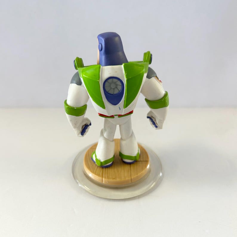 Puede incluir: Una figura de Buzz Lightyear, vista desde atr&aacute;s, se encuentra sobre una base circular transparente. El juguete presenta un traje espacial blanco y verde con un casco morado. La figura es un objeto de colecci&oacute;n de la franquicia Toy Story.