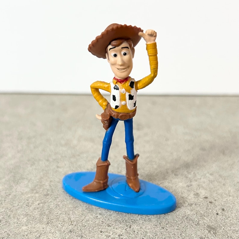 May include: A plastic figurine of a cowboy character wearing a brown hat, a yellow long-sleeve shirt, a white vest with black cow-print spots, blue pants, and brown boots. The figure stands on a blue oval base with one arm raised to the hat.