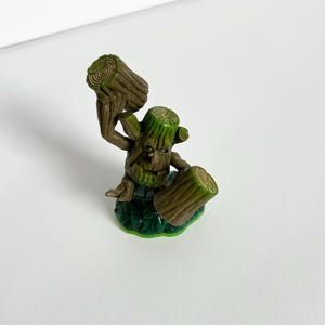 May include: A green and brown toy figure resembling a tree-like creature. The figure has a detailed wooden texture, holding two log-shaped objects. It stands on a green base with leaf-like details. The figure has a stern expression.