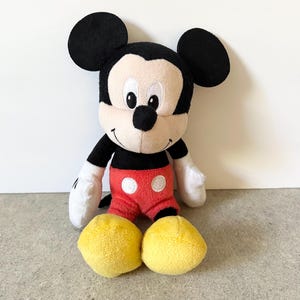 May include: A plush Mickey Mouse toy featuring large black ears, a cream-colored face with embroidered eyes, a black nose, and a wide smile. The character wears a black top, red shorts with two white circular buttons, white gloves, and yellow shoes.