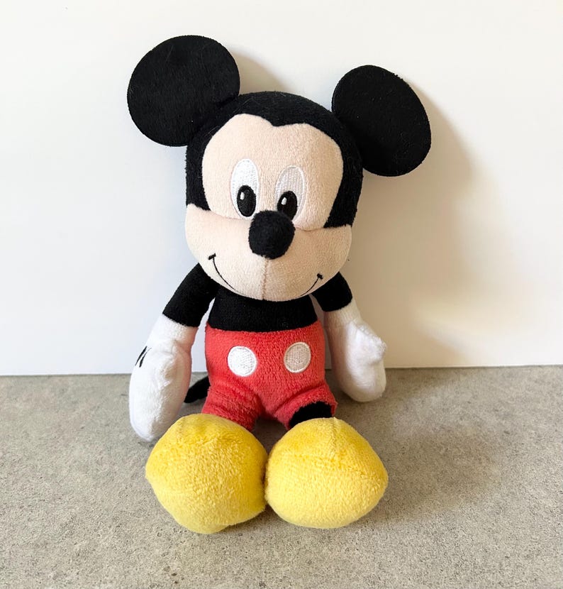 May include: A plush Mickey Mouse toy featuring large black ears, a cream-colored face with embroidered eyes, a black nose, and a wide smile. The character wears a black top, red shorts with two white circular buttons, white gloves, and yellow shoes.