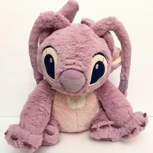 May include: A plush toy of the character Angel from the Lilo & Stitch franchise. The toy is primarily lavender with a light pink belly and inner ears. It has large, dark blue eyes and long, floppy ears.