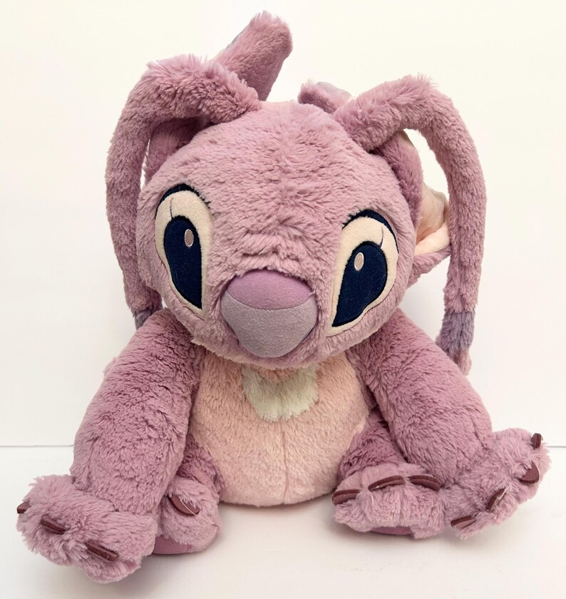 May include: A plush toy of the character Angel from the Lilo & Stitch franchise. The toy is primarily lavender with a light pink belly and inner ears. It has large, dark blue eyes and long, floppy ears.