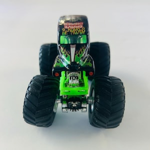 May include: A toy monster truck, the Grave Digger, is black with green flames and accents. The truck has large black tires and a green chassis. The name "Grave Digger" is printed on the top of the truck.