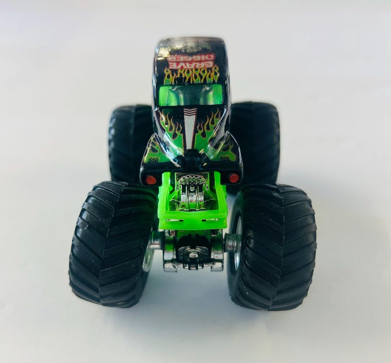 May include: A toy monster truck, the Grave Digger, is black with green flames and accents. The truck has large black tires and a green chassis. The name "Grave Digger" is printed on the top of the truck.
