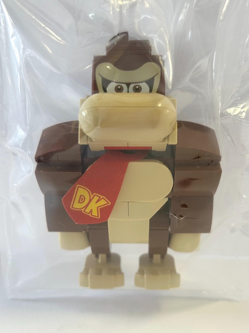 May include: A Donkey Kong figure made of brown, tan, and red building blocks. The figure has a red tie with "DK" printed on it. The figure is in a clear plastic bag.
