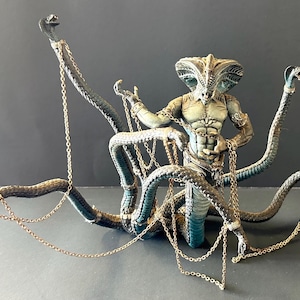 May include: A detailed figurine of a humanoid figure with a cobra-like head and multiple snake-like arms. The figure is painted in shades of green and tan, adorned with gold chains. The snakes are a dark teal color.