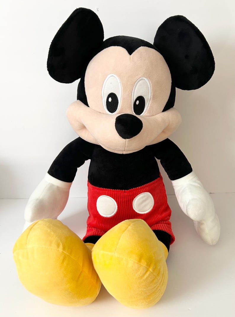 May include: A plush Mickey Mouse toy featuring large black ears, a tan face with white eyes, and a black nose. The character wears red corduroy shorts with two white circular buttons and large yellow shoes, with white gloves on its hands.