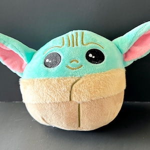 Pode incluir: Um brinquedo de pel&uacute;cia com um personagem com orelhas grandes, olhos pretos e um pequeno sorriso. O brinquedo tem uma cabe&ccedil;a e orelhas verde-claro com o interior das orelhas rosa. A parte inferior &eacute; castanha.