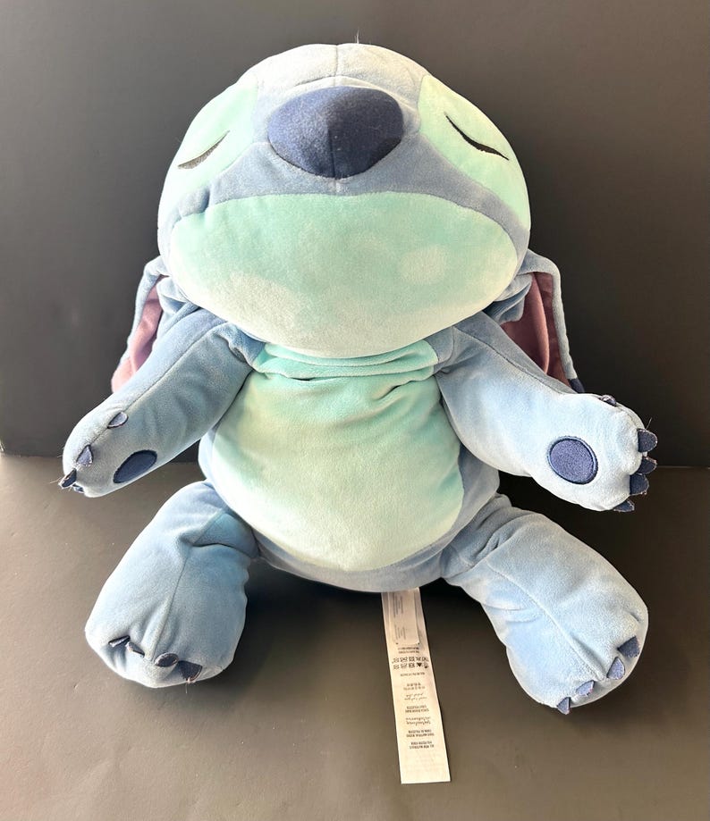 May include: A plush Stitch doll in shades of blue, green, and purple, with closed eyes. The soft toy features large ears, a round body, and a tag. The doll is designed to resemble the character from the animated film.